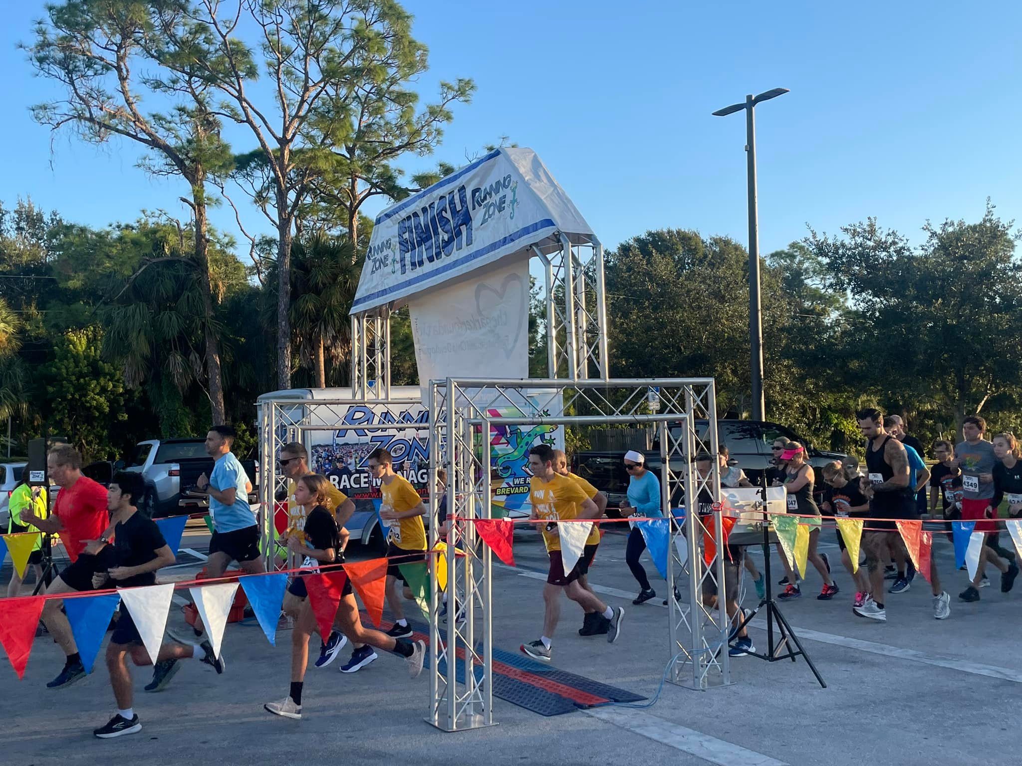 A candid shot of a group of runners as they cross the finish line of a 5k event. In the background are green, full trees and a blue cloudless sky.