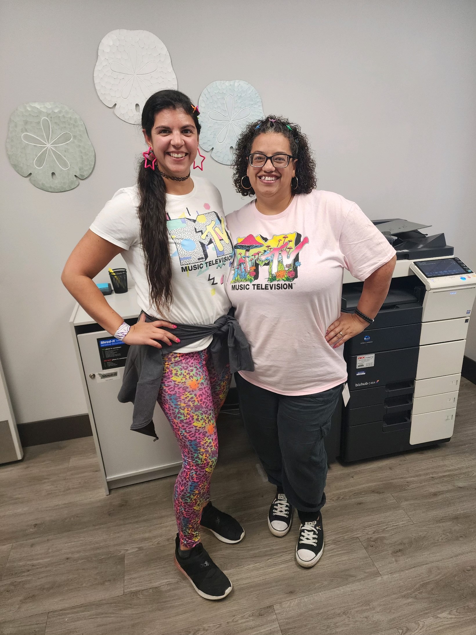 Two women in 90s outfits stand with their hands on their hip, smiling at the camera