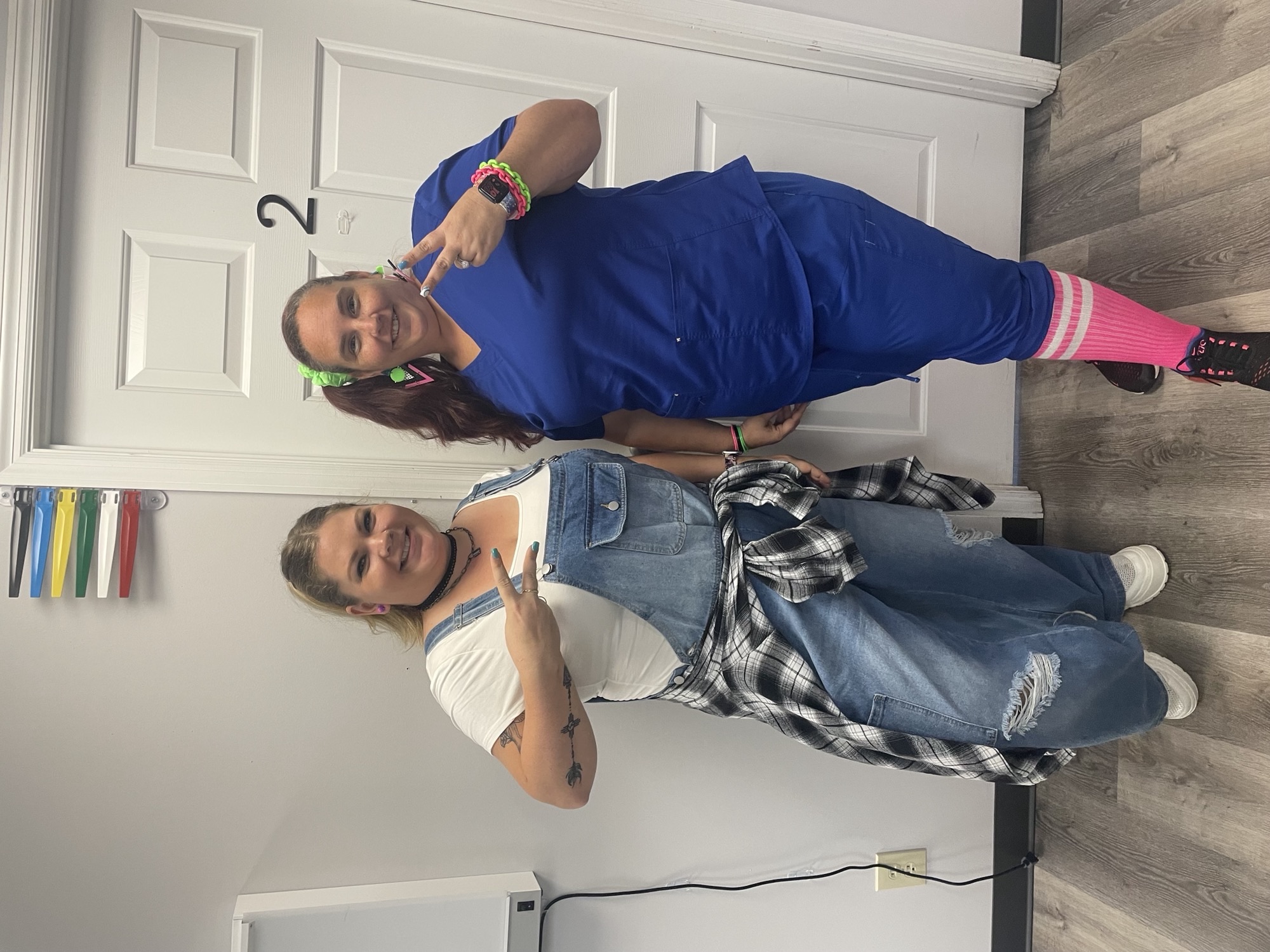 Two women dressed in 90s outfits make peace signs and smile at the camera