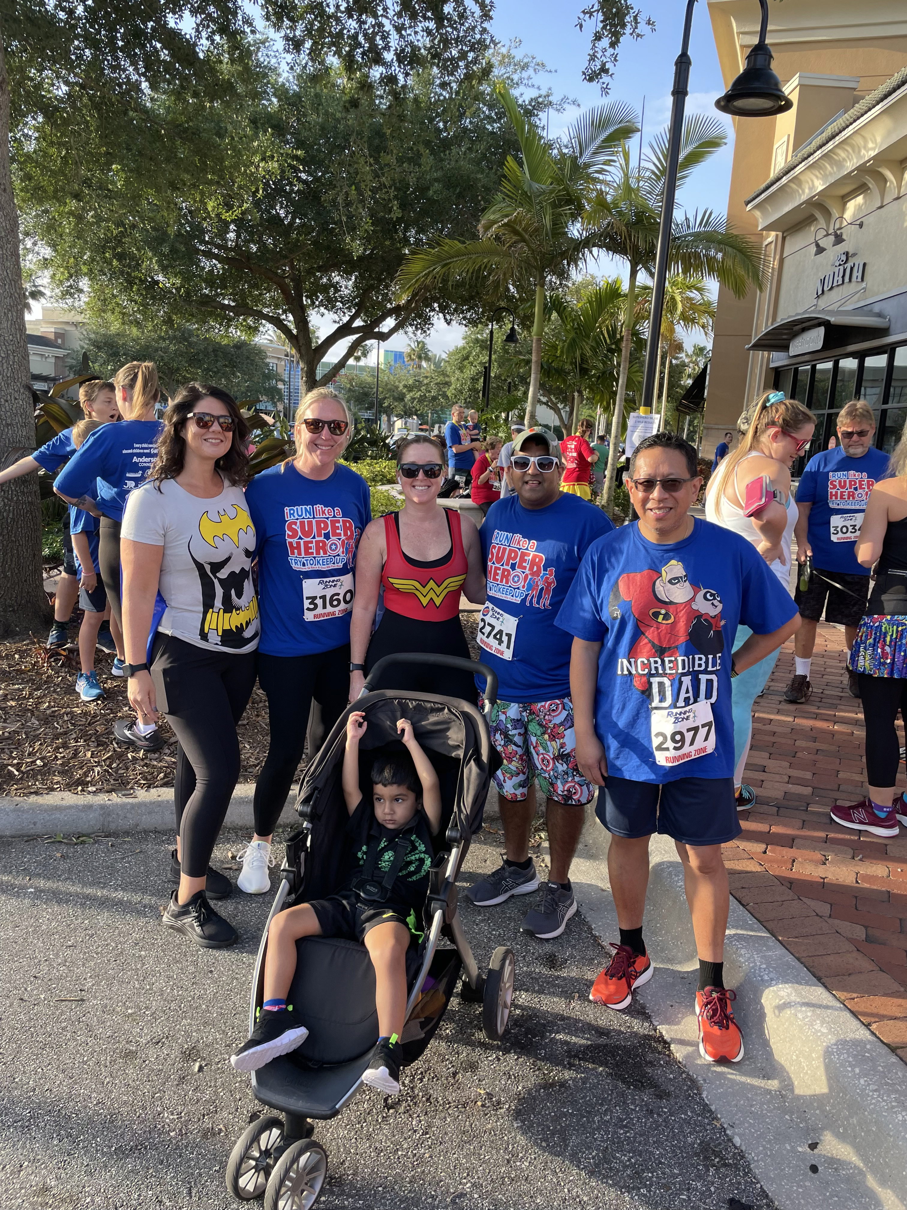 Group picture at charity event, five adults smiling at camera and child with arms raised in a running stroller